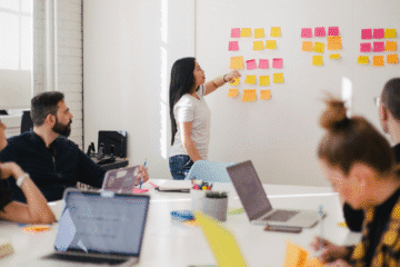 Pessoas sentadas ao redor de uma mesa, em que à frente de todas há uma mulher apontando para um quadro branco no qual estão fixados post-its