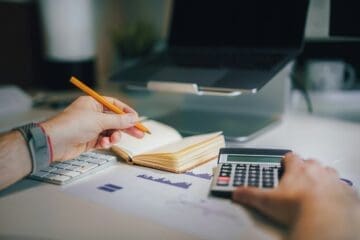 Mão esquerda de um homem segura um lápis laranja, já a direita, uma calculadora. Sobre a mesa estão: caderno, planilha de gastos, teclado e um notebook.